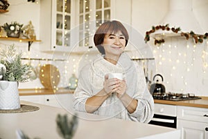 Happy young middle aged woman enjoying drinking tea or coffee in kitchen with Christmas decor