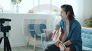 Happy young mixed race man playing guitar waving hand and talking recording video for online blog