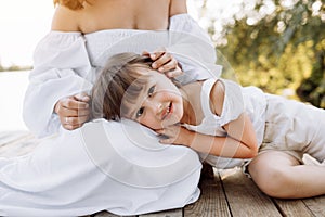 Happy young mom and child girl sitting near lake, pond. Mother and daughter smiling while spending free time outdoors