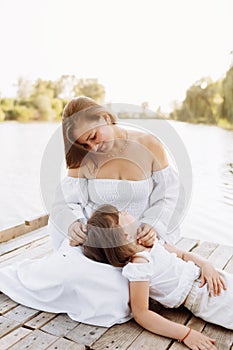 Happy young mom and child girl sitting near lake, pond. Mother and daughter smiling while spending free time outdoors