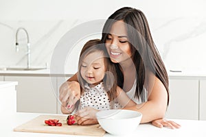 Happy young mom with little cute asian daughter cut strawberry