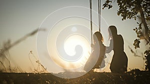 Happy young mom with a little girl swinging in the park together at sunset. Mother and daughter riding on swing, family