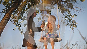 Happy young mom with a little girl swinging in the park together at sunset. Mother and daughter riding on swing, family