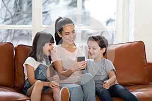 Happy young mommy sitting with children siblings, using mobile phone.