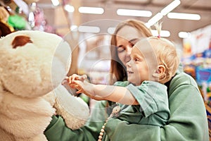 Happy young mother with cute baby in her hands walking around toy store in shopping mall. Mom and little blonde daughter