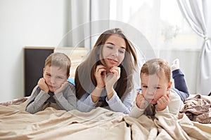 Happy young mother dressed in light blue pajama lays with her two little sons holding their hands under their chins on