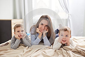 Happy young mother dressed in light blue pajama lays with her two little sons holding their hands under their chins on