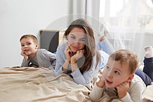 Happy young mother dressed in light blue pajama lays with her two little sons holding their hands under their chins on