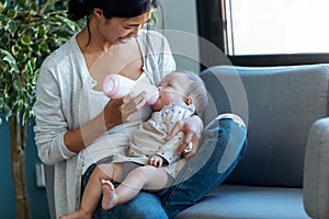 Happy young mother feeding her baby daughter with feeding bottle while sitting on sofa at home