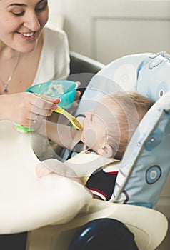 Happy young mother feeding her baby in highchair from spoon