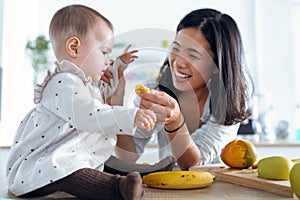 Happy young mother feeding her cute baby girl with a banana in the kitchen at home