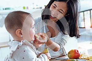 Happy young mother feeding her cute baby girl with a banana in the kitchen at home