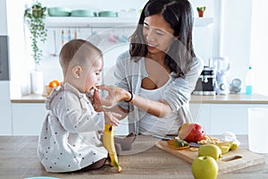 Happy young mother feeding her cute baby girl with a banana in the kitchen at home