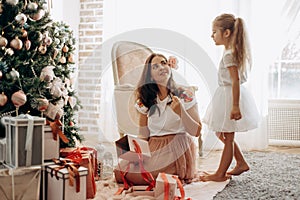 Happy young mother with flower in her hair and her little daughter in nice dress sit near the New Year`s tree and open