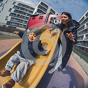 Happy young mother with her baby boy playing in colorful playground for kids. Mom with toddler having fun during