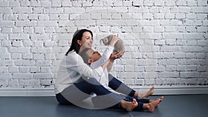 Happy young mother and her baby son kissing and hugging, playing with cat, resting at home near big window together