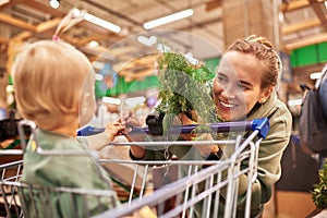 Happy young mother with her cute daughter in shopping trolley walking around supermarket. Beautiful mom and little