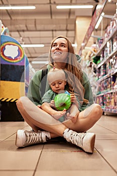 Happy young mother with her cute daughter walking around toy store in shopping mall. Mom and little blonde baby girl