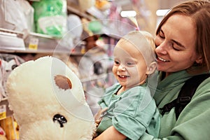 Happy young mother with her cute daughter walking around toy store in shopping mall. Mom and little blonde baby girl