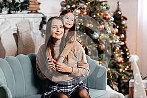 A happy young mother and her daughter are hugging in front of a beautifully decorated Christmas tree