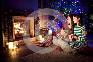 Happy young mother and her daughters having a good time sitting together by a fireplace in a cozy dark living room on Christmas