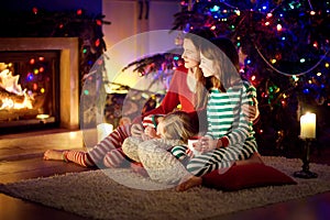 Happy young mother and her daughters having a good time sitting together by a fireplace in a cozy dark living room on Christmas