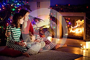 Happy young mother and her daughters having a good time sitting together by a fireplace in a cozy dark living room on Christmas