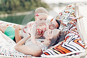 Happy young mother holding baby daughter in hands while relaxing in hammock with husband, hipster family concept, cute girl with