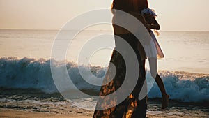 Happy young mother holding and spinning little daughter smiling at epic golden sunset beach, concept of family travel.