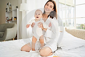 A happy young mother is hugging her newborn son while sitting on a large white bed