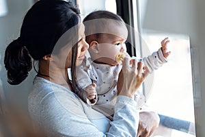 Happy young mother with little baby girl looking through the window at home