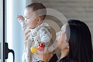 Happy young mother with little baby girl looking through the window at home