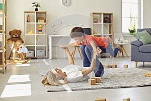 Happy young mother lying on carpet, holding her son on legs in raised hands and having fun at home