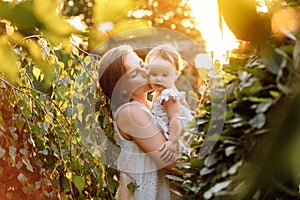 Happy young mother playing and kissing her little baby daughter on sunshine warm spring or summer day. Beautiful sunset