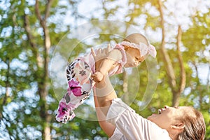 Happy young mother playing with little 5 months daughter in park. Lovely baby girl laughing while mother holding her in the air