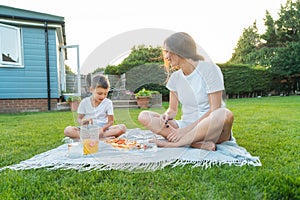 Happy Young mother and preschooler son having outdoor picnic dinner, eating pizza sitting on backyard Lawn on Sunny Day