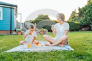 Happy Young mother and preschooler son having outdoor picnic dinner, eating pizza sitting on backyard Lawn on Sunny Day