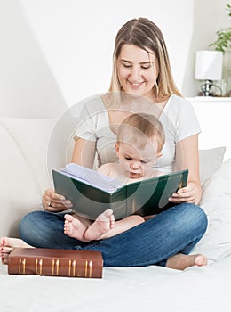 Happy young mother reading big old book to her baby boy ion bed