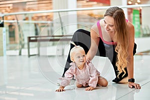 Happy young mother watch her cute baby crawling on floor at public place, shopping mall. Mom and her little daughter in