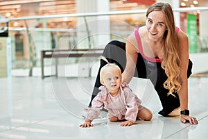 Happy young mother watch her cute baby crawling on floor at public place, shopping mall. Mom and her little daughter in