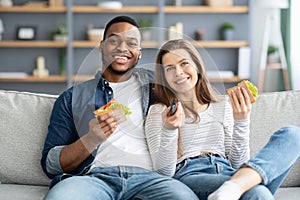 Happy Young Multicultural Couple Watching Tv And Eating Sandwiches At Home