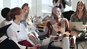 Happy young multiethnic business women sit together listening to corporate seminar laughing at modern office conference.