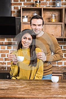 happy young multiethnic couple embracing and smiling at camera while drinking coffee