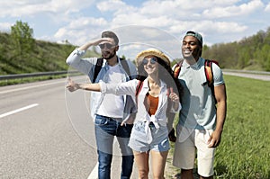 Happy young multiracial friends standing on highway, showing thumb up, hitchhiking for car, going on journey together