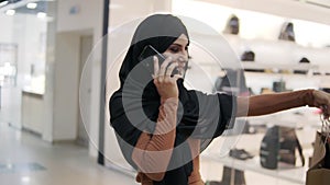 Happy young muslim woman in black hijab with professional makeup talking on smartphone walking by modern shopping mall