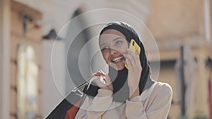 Happy young muslim woman in hijab talking on smartphone walking down the street with shopping bags in her hand.