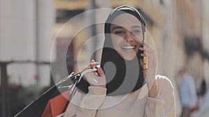 Happy young muslim woman in hijab talking on smartphone walking down the street with shopping bags in her hand.