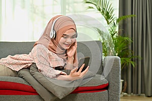 Happy young muslim woman listening to music on wireless headphone and using mobile phone in living room