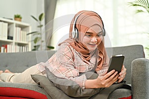 Happy young Muslim woman lying on couch and reading a text message on her mobile phone