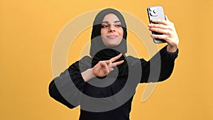 Happy young Muslim woman taking selfie by modern smartphone, portrait in orange background in studio